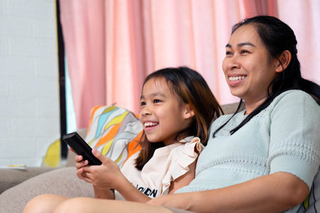 Happy Asian mother and daughter sitting on the sofa and watching TV together at home. The image...