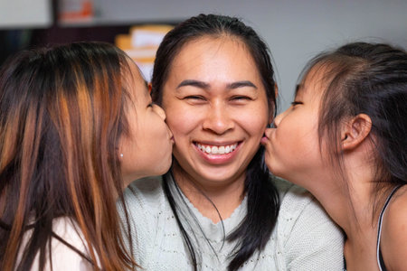 A joyful Asian mother smiling while her two daughters kiss her on both cheeks at home. The image represents family love, affection, motherhood, happiness, and bonding between parent and children. Perfect for illustrating Mother's Day, parenting, and emotional family moments.の写真素材
