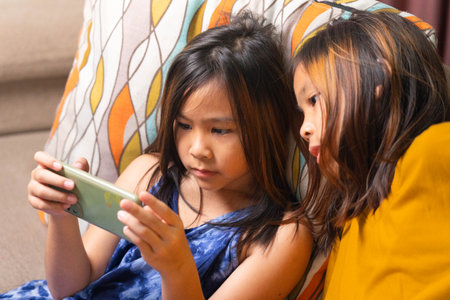 Two Asian girls sitting on sofa watching smartphone screen together at home, representing childhood, learning, technology, and digital lifestyle in modern family life.の写真素材