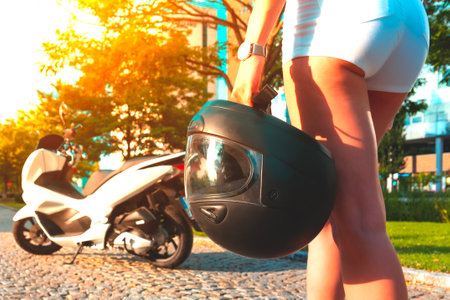 Girl in white shorts with black motorcycle helmet walks to the white motorcycle. Young woman with moto scooter at sunny day. Back view.の写真素材