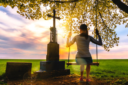 Young woman on a wooden swing hanging on a tree at monument with a Christian cross at sunset. Young people and religion.の写真素材