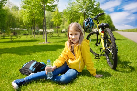 Teenage girl biker sitting on a green grass next to a mountain bike and drinking waterの写真素材