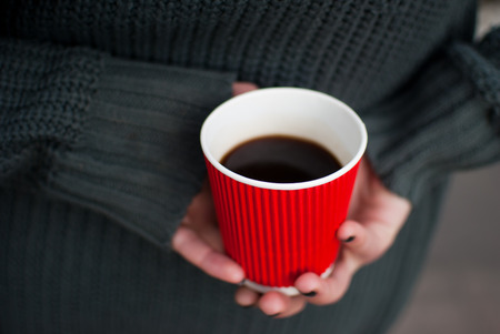 The thin girl dressed in a grey sweater holds a red paper cup of coffee in her pale beautiful hands.の写真素材