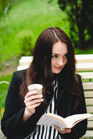 Smiling brunette girl sitting on a bench in a park and reading a synopsis of a soft-cover book.の写真素材