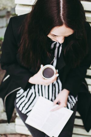 Smiling brunette girl sitting on a bench in a park and reading a soft-cover book.の写真素材