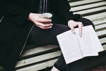 Young girl holding an open book and paper cup of coffee in her hadns while seated on a bench in a...