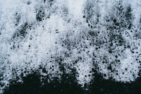 White ocean water foam on the black sand covered with small stonesの写真素材