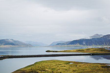 Colorful Icelandic landscapes under gray heavy sky. Green wet grass, beautiful lake, and snowy tops of the mountais reflected in the lake water. Kirkjufellsfoss, Snaefellsnes peninsula.の写真素材
