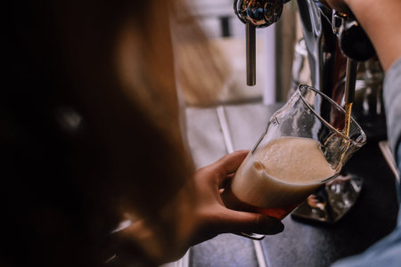 Girl pouring a pint of dark beer in a craft beer pub. Local pub. Shiny beer taps on the background. Bright sunny afternoon. Look from a shoulderの写真素材