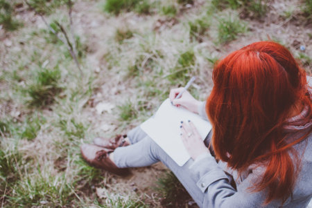 Redhead girl wearing a grey coat sitting on a green spring grass writing down her thoughts in the notebook. Pale hands and blank paper of the notebook. Concept (idea) of thinking, dreaming, planning, learning, and studying. Early spring afternoon.の写真素材