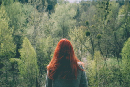 Longhair redhead girl wearing a grey coat standing on the edge of the hill in front of the forest of...