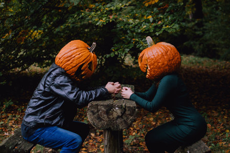 A couple, with pumpkins on their heads, sitting behind a wooden table in an autumn forest. Halloween costume idea for a coupleの写真素材