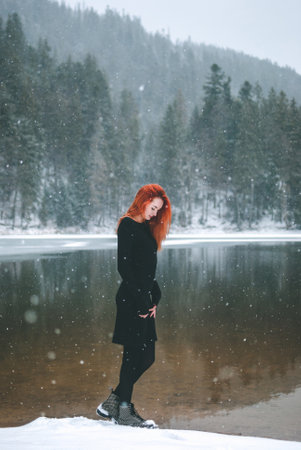 Handsome redhead girl looking down. Snowflakes falling on her hair. Splendid shady black mountain lake. Dreamy winter landscape of Carpathian mountains.の写真素材