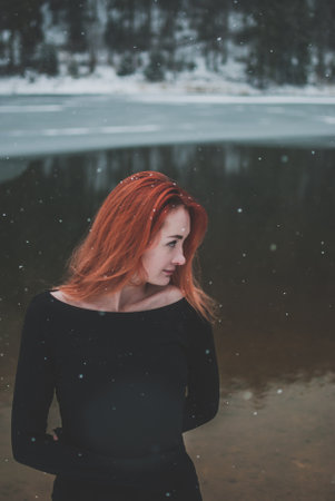 Handsome redhead girl looking in the distance. Snowflakes falling on her hair. Splendid shady black mountain lake. Dreamy winter landscape of Carpathian mountains.の写真素材