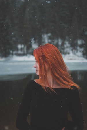 Handsome redhead girl looking in the distance. Snowflakes falling on her hair. Splendid shady black mountain lake. Dreamy winter landscape of Carpathian mountains.の写真素材