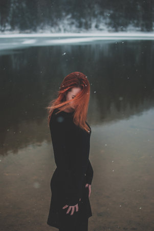 Handsome redhead girl looking down. Snowflakes falling on her hair. Splendid shady black mountain lake. Dreamy winter landscape of Carpathian mountains.の写真素材
