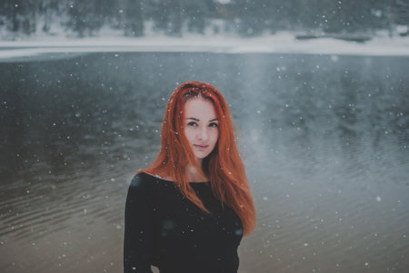 Joyful handsome black-eyed redhead girl looking at the camera. Splendid shady black mountain lake reflecting trees of forest behind her back.の写真素材