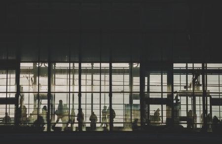 Black sillhouettes of people waiting for their train on a subway station behind glass windows filled...