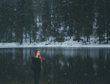 Snowflakes flying over handsome black-eyed redhead girl looking in the distance. White snowflakes flying all around. Splendid shady black mountain lakeの写真素材