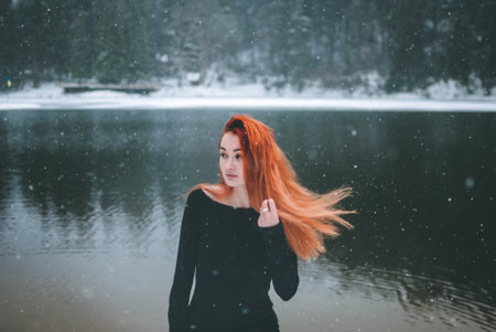 Joyful black-eyed redhead girl looking in the distance. Splendid shady black mountain lake reflecting trees of forest behind her back.の写真素材