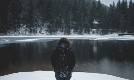 Snowflakes flying over long-haired man looking in the distance and standing by splendid shady black mountain lake.の写真素材