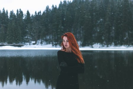 Joyful handsome black-eyed redhead girl looking in the distance. Splendid shady black mountain lake reflecting trees of forest behind her back.の写真素材