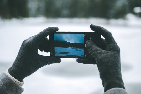 Gilr taking a photo of a beautiful mountain lake Synevyr in Ukraine on her black smartphone. Frozen lake covered with snow against dark winter forest and mountains covered with clouds. Gray cloth glovesの写真素材