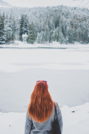 Redhead girl looking in the distance. Frozen winter lake covered with ice and snow. Snow-white fir trees of the forests on the background. Winter landscape in the Carpathian mountainsの写真素材