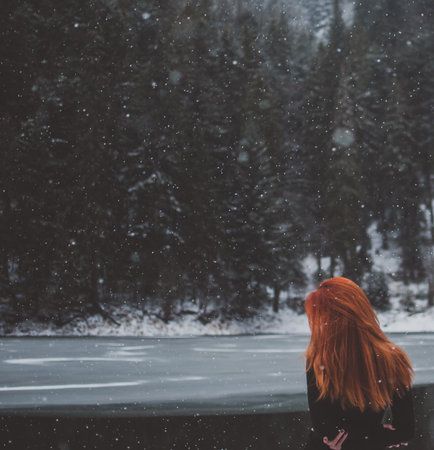 Square-shaped. Handsome redhead girl looking in the distance. Snowflakes falling on her hair. Splendid shady black mountain lake. Dreamy winter landscape of Carpathian mountains. Look from behindの写真素材