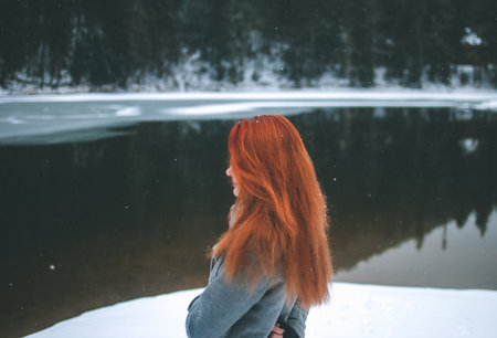 Handsome redhead girl looking in the distance. Splendid shady black mountain lake reflecting trees of forest behind her. Dreamy winter landscape of Carpathian mountainsの写真素材