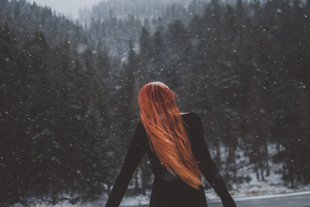Handsome dark-eyed redhead girl pretending she's flying by mountain lake called Synevyr and looking up in the distance. Snowflakes descending on her beautiful hair. Concept of romance, freedom, happiness, and mental health.の写真素材