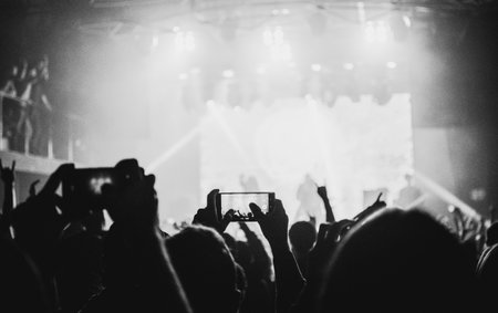 Black and white photograph of a concert audience with raised hands, smartphones recording, stage lights, and a vibrant atmosphere of energy and freedom.の写真素材