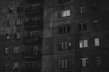 Black and white photo of a night-time socialist-era apartment block with lit windows, evoking solitude and urban alienation.の写真素材