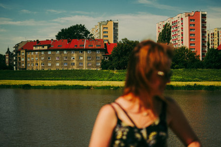 A woman with red hair stands by the riverbank in Opole, Poland, with blocks of socialist architecture in the background and nostalgic, film-like tones evoking a retro mood.の写真素材