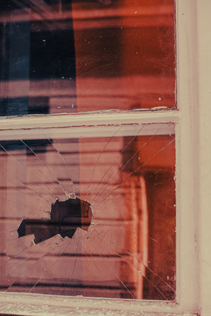 A broken windowpane with spiderweb-like cracks and warm-toned reflections. The scene evokes a sense of abandonment, imperfection, and quiet tension.の写真素材