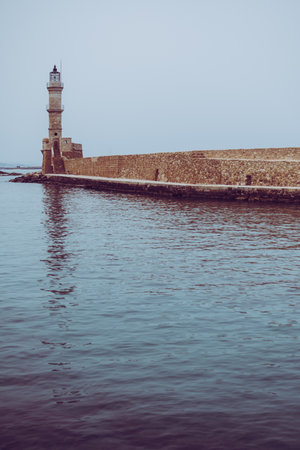 The historic lighthouse of Chania stands at the edge of the Venetian harbor in Crete, Greece, bordered by calm Mediterranean waters and a stone walkwayの写真素材