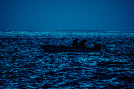 Silhouetted fishermen with fishing rods on a boat drift across dark blue water at dusk. A calm yet dramatic seascape that conveys solitude, friendship, and life at sea.の写真素材