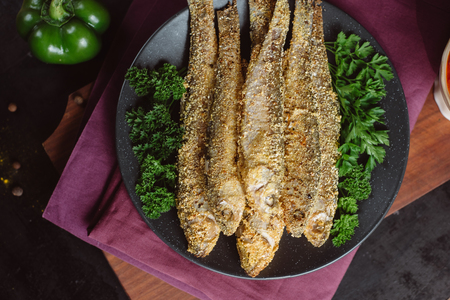 Fried fish in a plate with tomato sauce, greens and peppers on a wooden board and a dark background