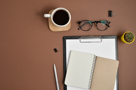 Mockup workspace with blank clip board, office supplies, cactus, pen, coffee cup, notepad and glasses on brown background. Flat lay, top view, copy spaceの写真素材