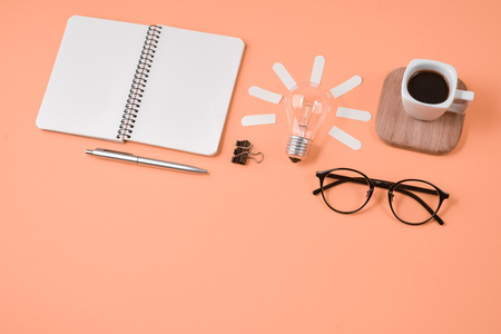 Top view flat lay of workspace desk styled design office supplies with pen, notepad, eyeglasses, cup coffee and light bulb on a orange color paper background minimal style. Concept brainstormingの写真素材