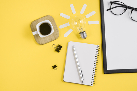 Top view flat lay of workspace desk styled design office supplies with pen, notepad, eyeglasses, cup coffee and light bulb on a yellow color paper background minimal style. Concept brainstormingの写真素材