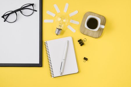 Creative flat lay concept top view of workspace desk styled design office supplies with pen, notepad, eyeglasses, cup coffee and light bulb on a yellow background. Concept brainstormingの写真素材