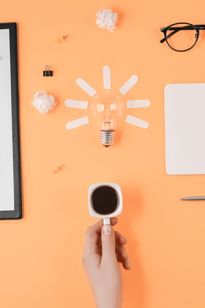 Flat lay brainstorming table top view. Blank clip board, office supplies, pen, notepad, eyeglasses, cup of coffee in hand, light bulb on orange background. Concept new ideaの写真素材