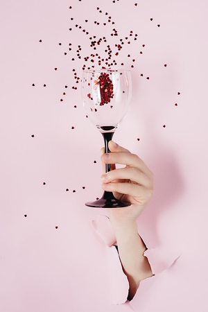Close up of female hand holds wine glass is seen through a hole in torn pink paper with splash of red heart shaped confetti. Overhead view, copy space. Holiday and birthday concept.の写真素材