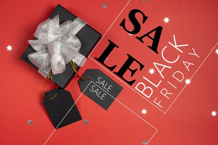 Gift box wrapped in black paper with silver ribbon price tags, stars confetti and text on bright red background. Copy space and top view. Black friday box gift present isolated.の写真素材