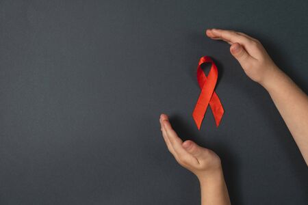 Top view on child's hands holding red ribbon on dark background, HIV awareness concept, world AIDS...