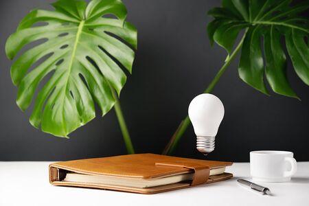 Close up of home office desk table workspace with a notebook, light bulb, pen, cup coffee, green leaf and eyeglasses on red background. Copy space mockup template.の写真素材
