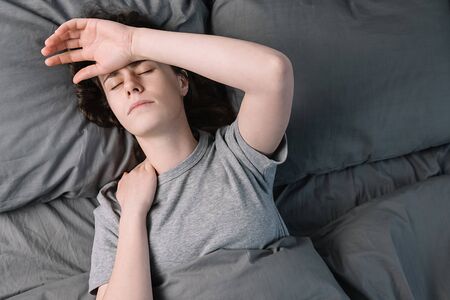 Top view on unhappy young woman with closed eyes lying in bed, touching forehead hand having a...