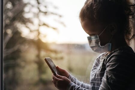 Tired little girl in medical sterile face mask sitting on window sill using modern smartphone gadget...