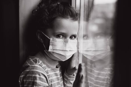 Black and white portrait of little girl sit on window sill wears virus mask, thinks abot risk of epidemic disease, child looks sadly at camera. Concept health and safety, coronavirus, virus protectionの写真素材
